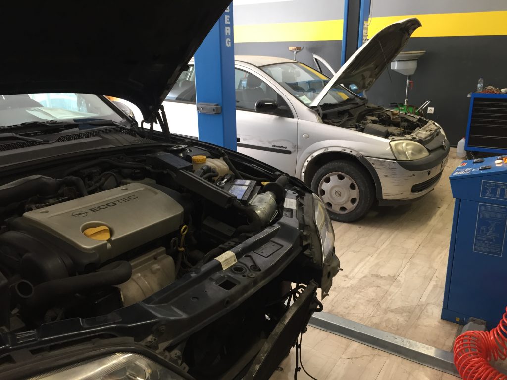 Ανοιχτός κινητήρας σε πρώτο πλάνο και Opel Corsa σε ανυψωτικό στο βάθος